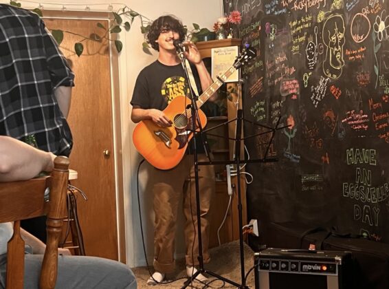 A person smiles at a mic stand, holding a guitar and getting ready to perform for a room of people.